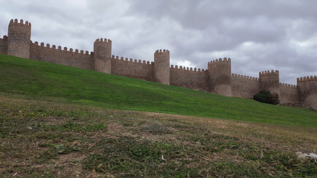 Ampia veduta delle storiche mura medievali di Ávila, Spagna, che si estendono su una collina erbosa sotto un cielo nuvoloso e drammatico, una delle città fortificate meglio conservate d'Europa e un sito patrimonio UNESCO