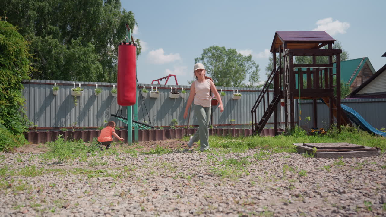White Woman Training With Punching Bag In Backyard, Child Nearby Exploring Playset, Fitness Session Under Clear Sky, Gloves, Swing, Gravel Ground, Energetic Motion, Casual Clothing