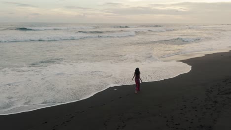 Mujer-Vestida-De-Rojo-Caminando-Por-La-Playa-De-Arena-Negra-En-El-Paredón,-Guatemala-Al-Atardecer---Toma-Aérea-De-Drones