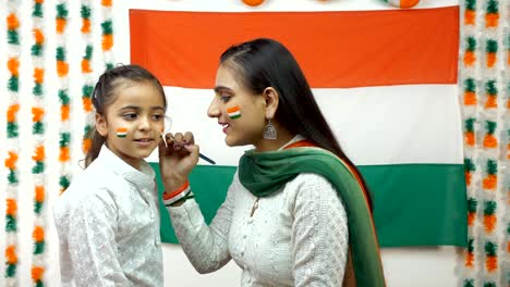 indian woman painting the face of a little girl in tricolors of a national flag - independence day / republic day concept