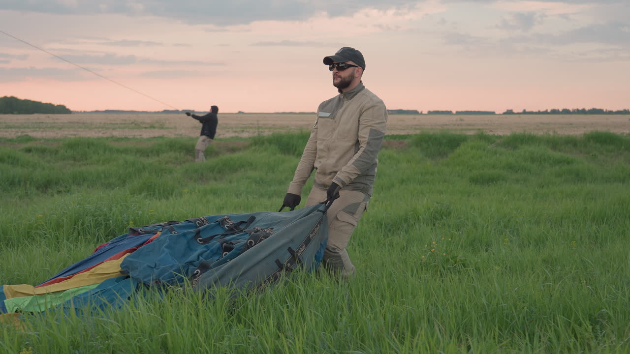 man in cap and gloves dragging heavy deflated hot air balloon envelope gear across grassy field at sunset during preflight pack up sequence with support crew securing ropes for safe landing