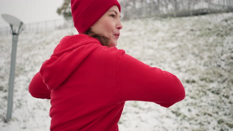 woman performs outdoor workout with hands up for balance, turning left and right in snowy setting with a snowy hill, iron railing, and blurred light pole in background