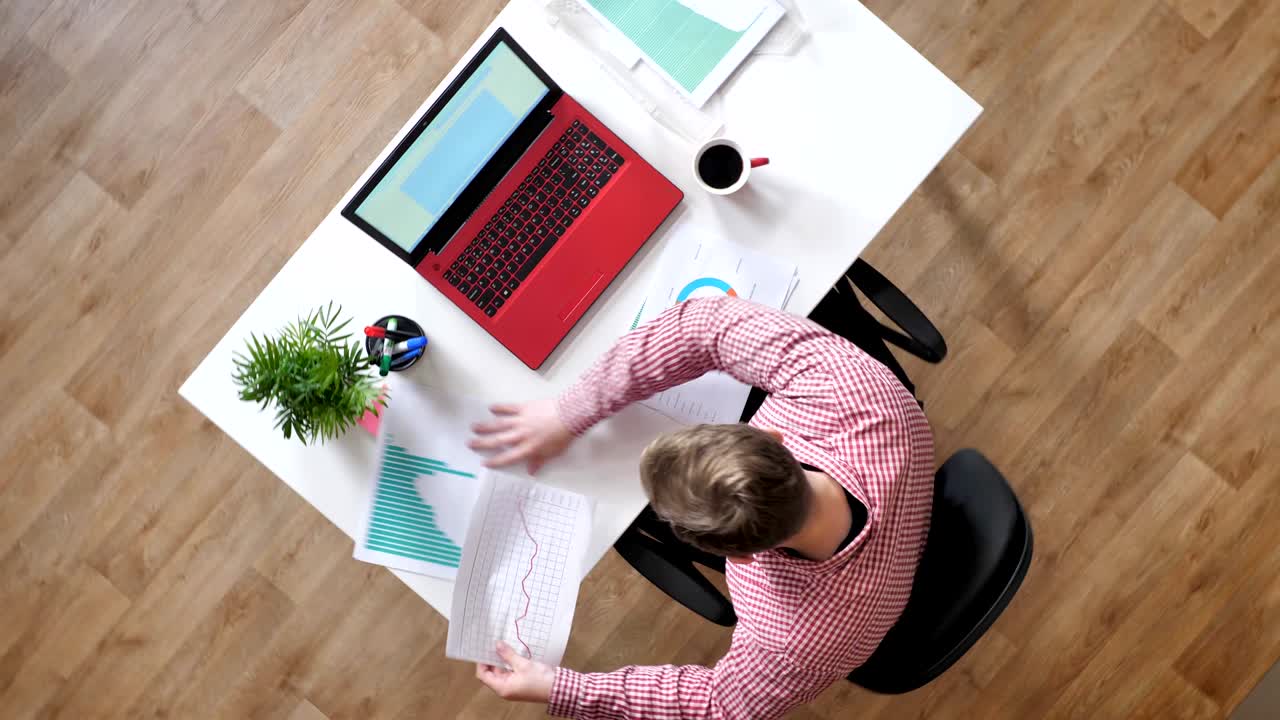 Young man in glasses typing on laptop, stressed and nerved, topshot, sitting behind desk with coffee, circling around view