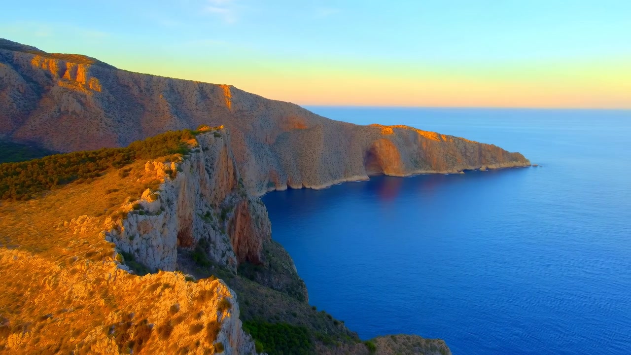Paesaggio costiero obliquo elevato che mostra un promontorio calcareo frastagliato, un altopiano roccioso, un'apertura di grotta, erba secca, arbusti sparsi e boschi sottostanti, mentre la calda luce del sole si sposta su pietra e acqua
