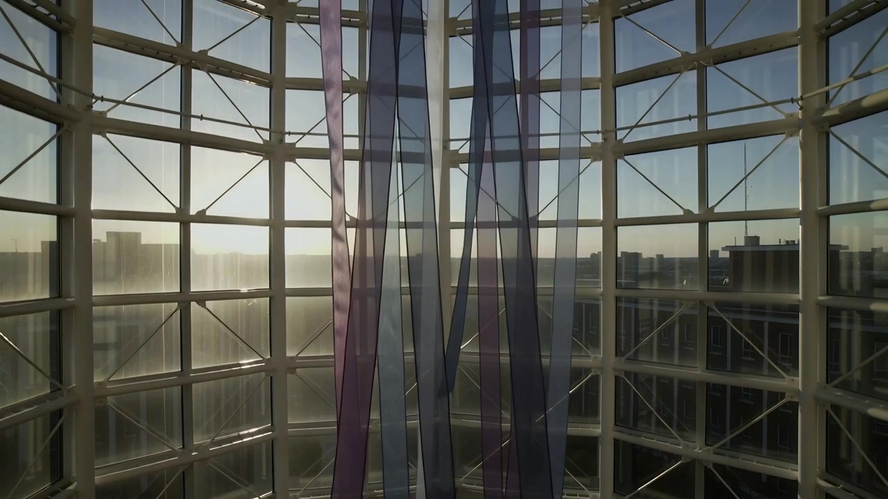 Swaying translucent purple-blue ribbons shifting in gentle breeze at curved glass wall, sun glare