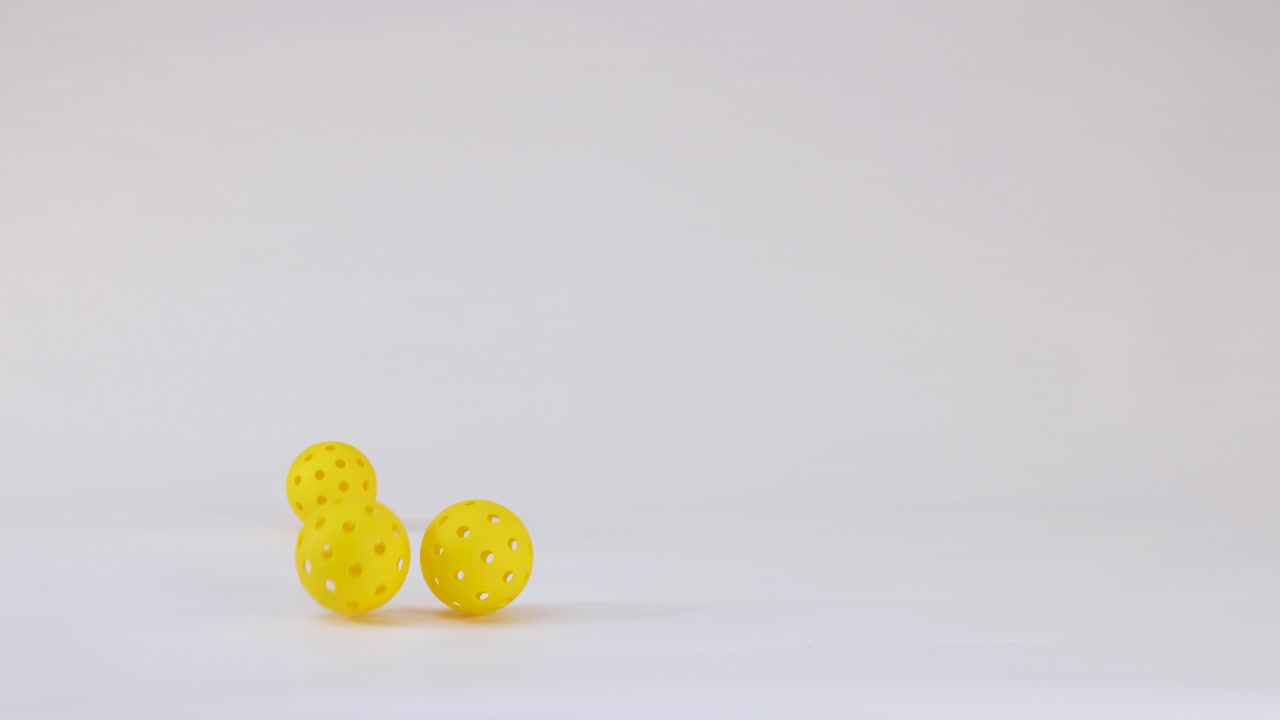 Three yellow plastic pickleball balls fall, bounce, and roll across a clean white studio background