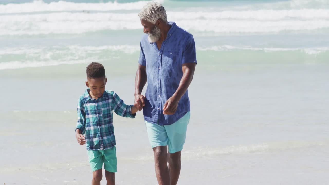 Happy senior african american father walking with son on sunny beach