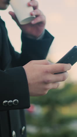 primer plano de un individuo con una bata negra haciendo varias tareas operando el teléfono y bebiendo té al aire libre, mostrando el enfoque y el estilo de vida moderno en un entorno urbano con un fondo borroso y un coche en movimiento