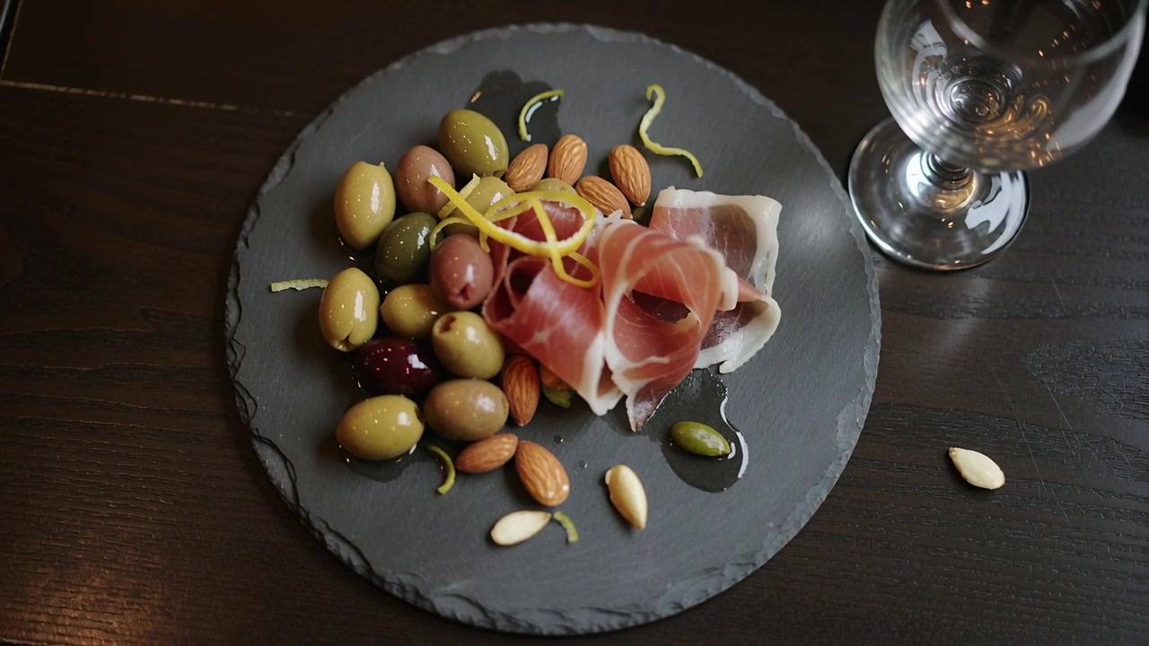 Kamera, yemek masasındaki başlangıç tabağına yaklaşarak prosciutto, zeytin ve yağın dokularını vurguluyor