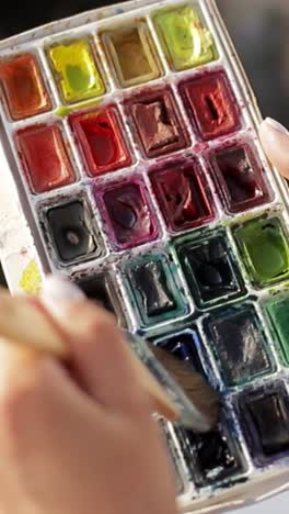 closeup of womans hand painting with brush on colorful palette a closeup shot of a womans hand adorned with beads delicately painting with a brush on a colorful palette the artist wears a white dress and the scene is bathed in soft natural light