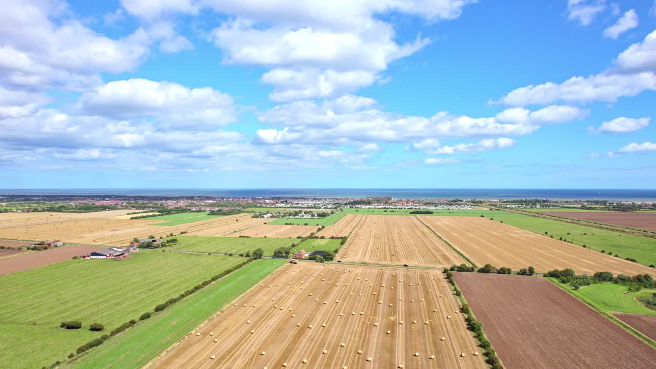 An aerial video frames the beauty of wind turbines in a row, gently spinning amidst a Lincolnshire farmer's freshly harvested field, adorned with golden hay bales