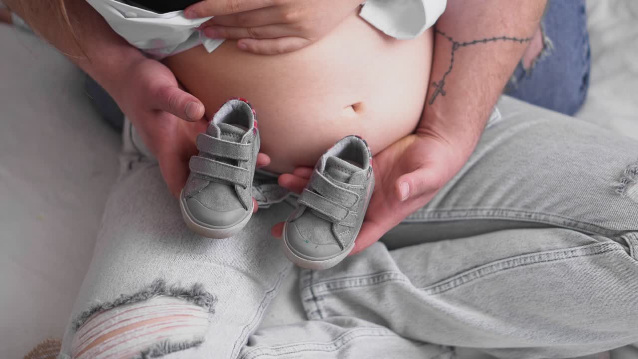 임신한 젊은 여성과 남편은 아기를 기대하고 있습니다. 젊은 가족은 보충을 기다리고 있습니다. 집에서 휴식을 취하는 큰 배를 가진 행복한 임신한 여성. 젊은 임신하는 어머니는 그녀의 배를 만져줍니다.