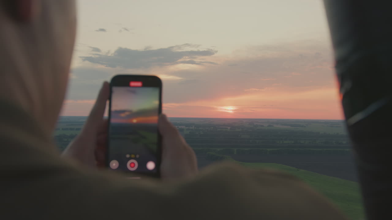 Rear view of lady recording breathtaking sunset using smartphone while floating in hot air balloon over vast countryside, glowing sky and layered farmland unfolding into distance