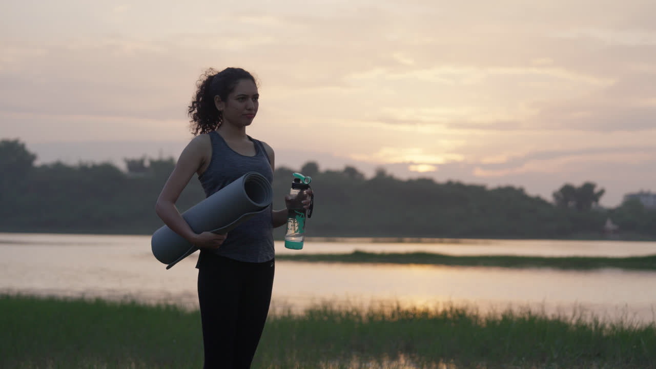 A wide-angle shot of a South Asian woman holding a yoga mat and water bottle beside a calm lakeside at sunrise, standing in soft warm morning light
