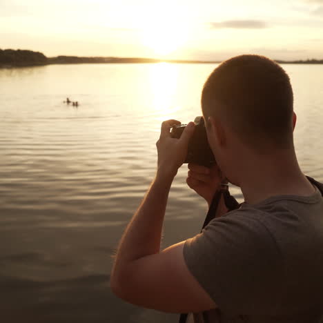a man is standing with his back and photographing the sunset on the lake surrounded by trees on the background with bathing people.