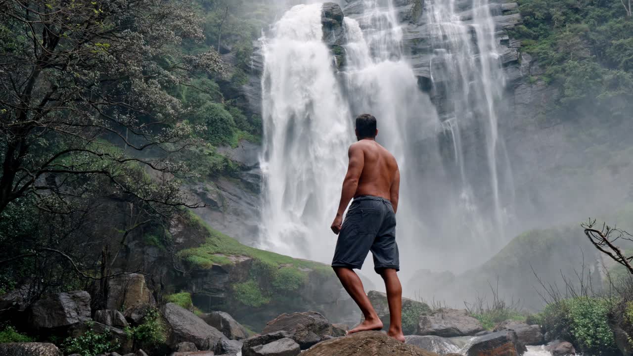 A man stands near the base of Bomburu Ella Waterfalls, surrounded by mist, rocks, and dense greenery in the highlands of Nuwara Eliya, Sri Lanka.
