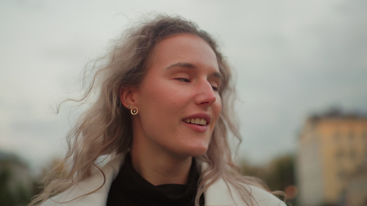 Portrait view of tourist with curly hair smiling and admiring surroundings, standing outdoors with blurred view of residential buildings, green trees, and soft evening sky in urban environment