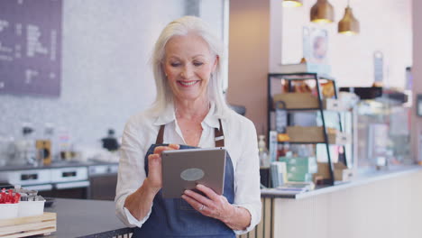 Portrait-Of-Mature-Female-Owner-Of-Coffee-Shop-Or-Restaurant-Using-Digital-Tablet