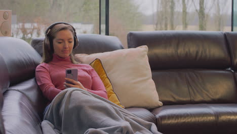 woman wearing wireless headphones listening to music streaming from mobile phone on sofa at home