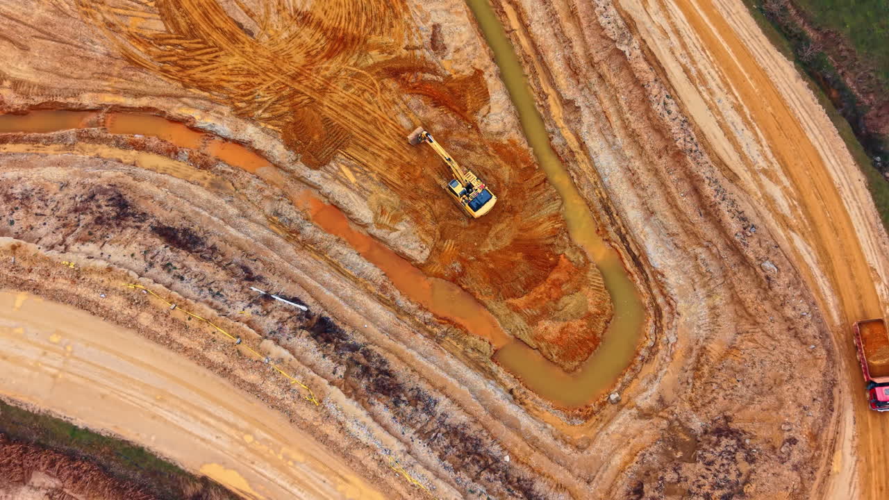 Top down view of yellow excavator working in sand pit. Detailed aerial shot of hydraulic shovel digging earth near water channel in open cast mine