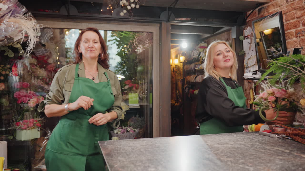 Due fioristi che sorridono al bancone del negozio, grembiuli verdi e fiori vivaci nella vetrina sullo sfondo, ritratti ravvicinati mostrano proprietari accoglienti pronti a servire, calda luce naturale e allestimenti artigianali.