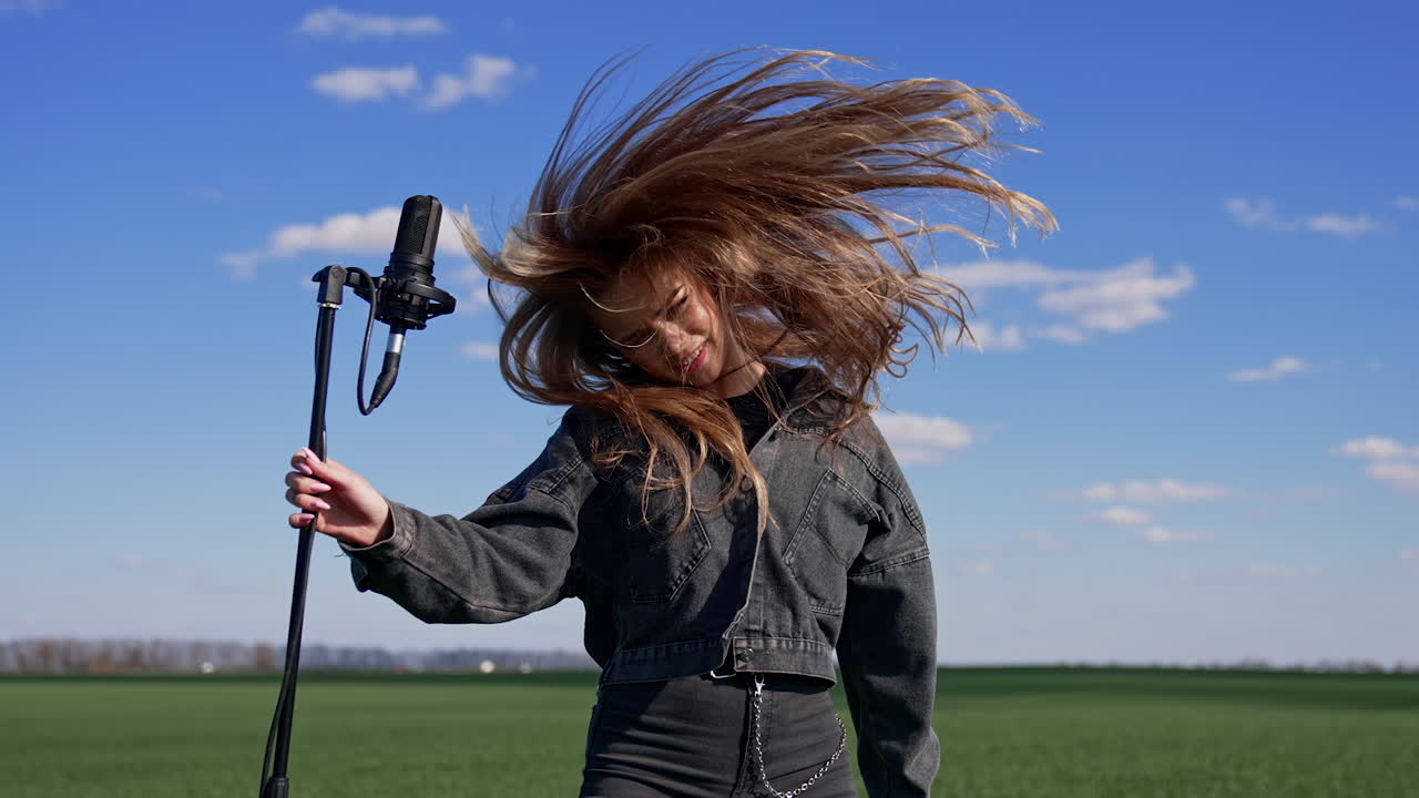 Pop female singer near the microphone outdoors. Pretty girl in denim suit twirling her head with long hair while singing on green field background.