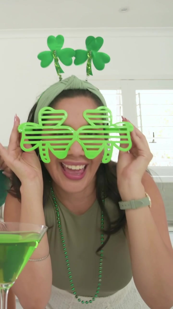 Vídeo vertical: Mulher sorridente de verde tirando os óculos de sol e bebendo uma bebida verde na cozinha para o Dia de São Patrício.