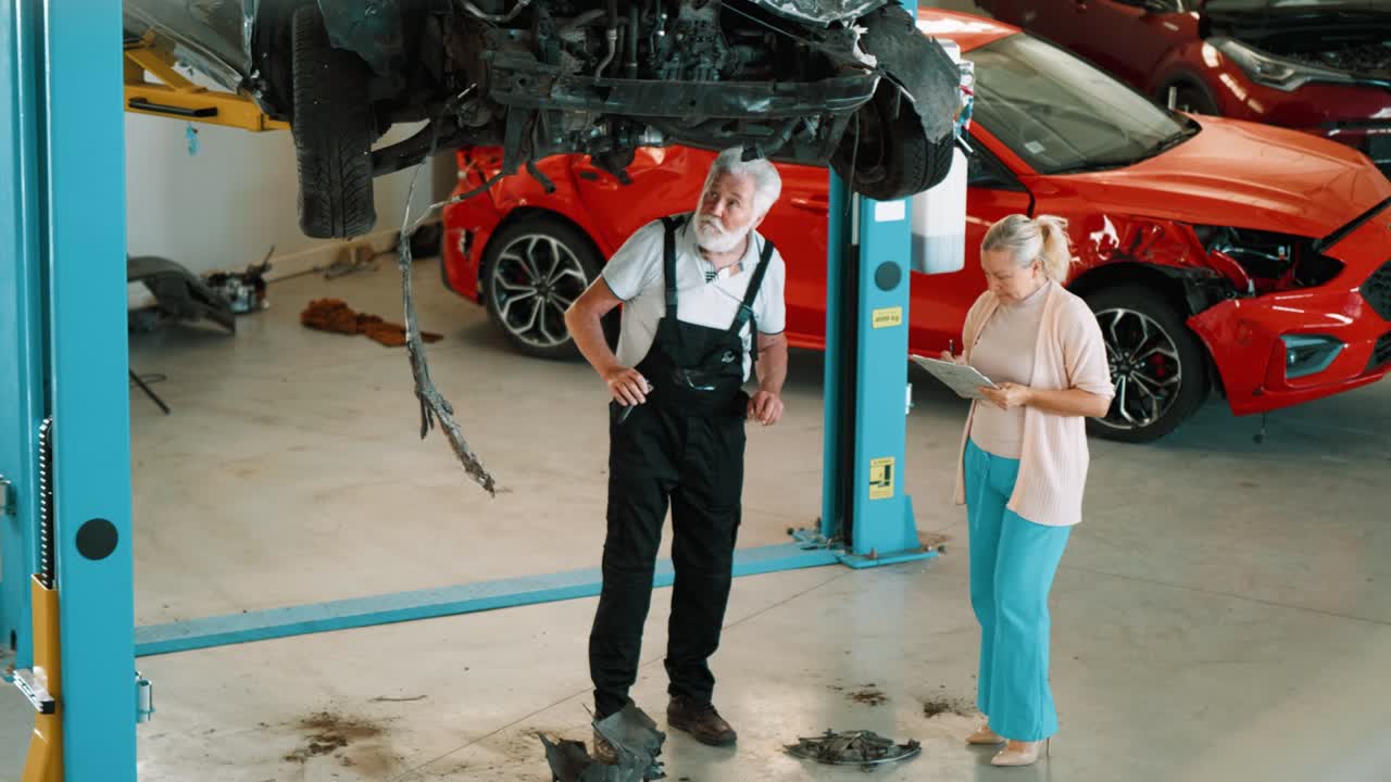 vista superior de una mujer adulta inspector de seguros y un hombre mecánico anciano en el taller inspeccionan un coche chocado levantado en una grúa
