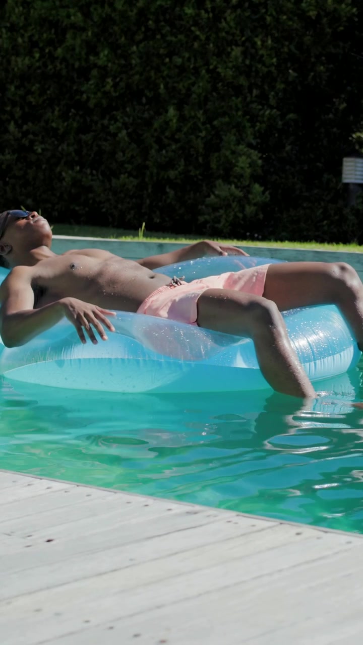 Vertical video: Lying man shifting weight, drifting on blue ring in yard pool in pink trunks shades