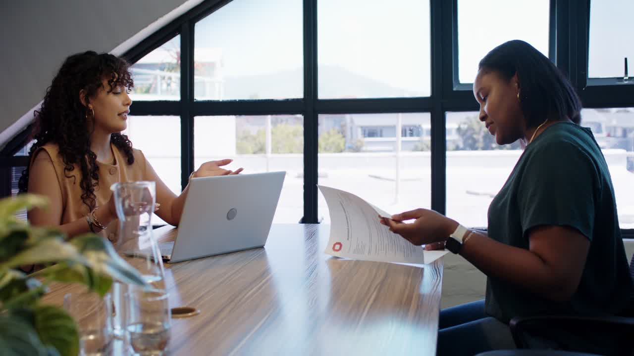 African American women colleagues in office at table, left using laptop, causing right reading docs