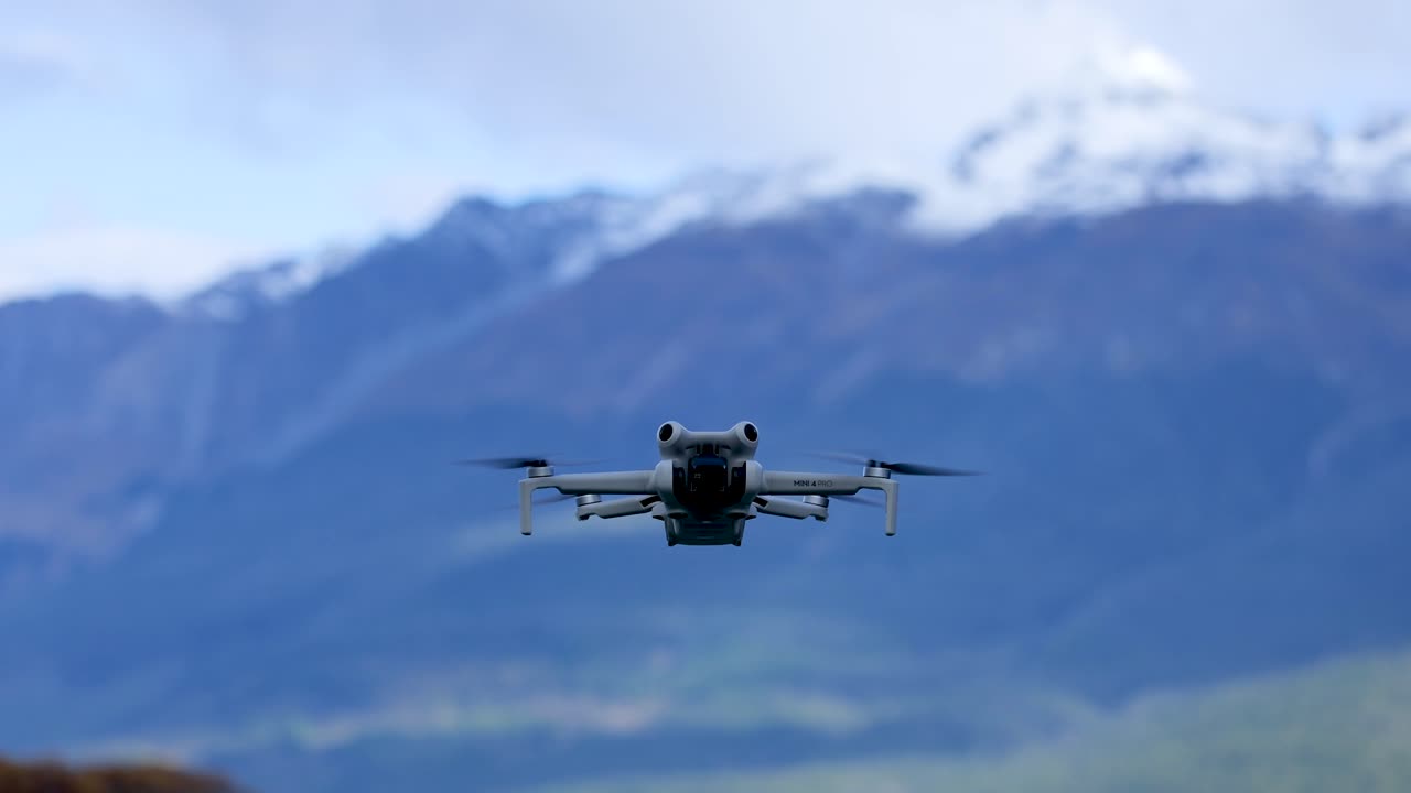 Quadcopter drone flies forward toward camera with snowy mountains and blue sky in background
