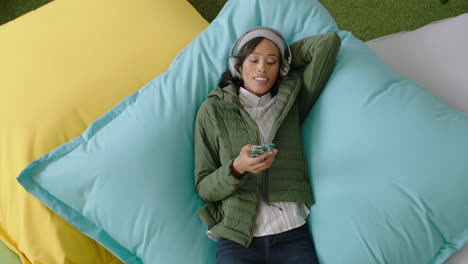 happy african american woman student using smartphone browsing online social media messages enjoying lunch break relaxing on colorful pillows listening to music in modern office top view