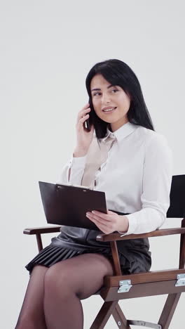 brunette girl talking the phone isolated in studio. young business woman in white blouse sitting in the chair with a folder and speaking the mobile phone. slow motion. vertical video