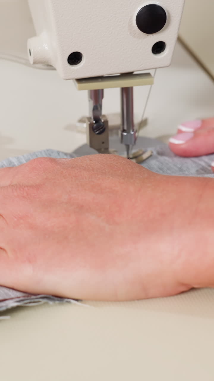 Manos caucásicas guiando tela vaquera bajo la aguja, primer plano de una máquina de coser industrial cosiendo un dobladillo en bruto, palmas firmes con uñas cuidadas, trabajo de reparación de precisión en un pequeño taller