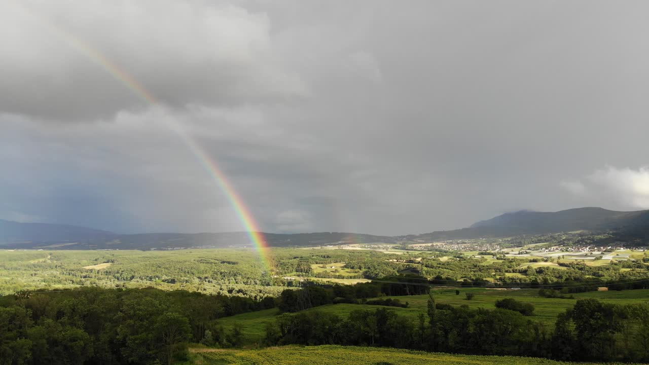 Aerial footage of a rural area in France featuring rolling hills, cultivated fields, a small village, and a vivid rainbow stretching across the sky.