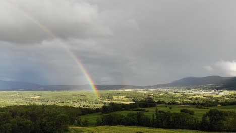 aerial footage of a rural area in france featuring rolling hills, cultivated fields, a small village, and a vivid rainbow stretching across the sky.