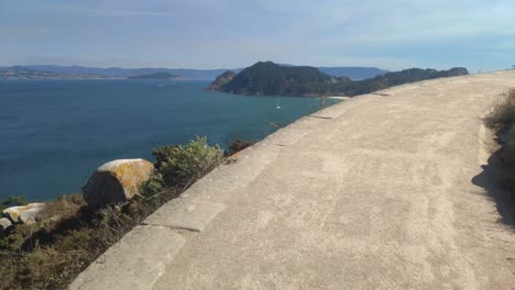 Going-up-the-road-with-trees,-mountainous-island-with-beach-and-sailboats-sailing-on-a-sunny-day,-panoramic-shot-traveling-forward-roll-to-the-right,-Cíes-Island,-Pontevedra,-Galicia,-Spain