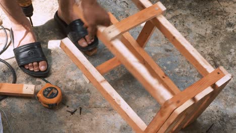 carpintero hábil que emplea un taladro eléctrico para colocar tornillos en una pequeña silla de madera en su pequeño taller para vender y apoyar la economía local
