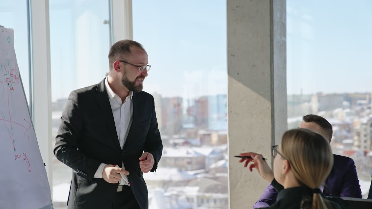 Positive and warm atmosphere at morning meeting. Smiling man makes speech about chart in front of his teammates. Sunlit cityscape at backdrop.