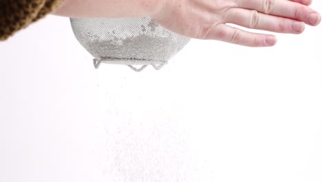 white female hand sifting powdered sugar in sieve on white backdrop in slow motion