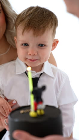 black cake with mickey mouse is held in front of a little caucasian baby boy. a candle is lit by mom. vertical video.