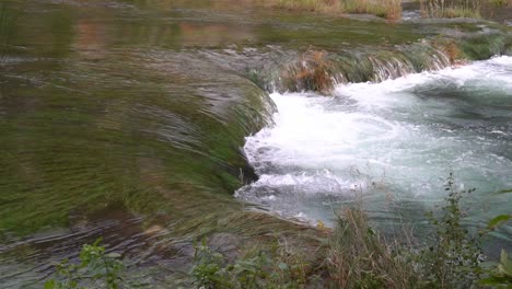 close up of a strong current of water flowing from one pond down to another submerging the grass nearby at ¼ speed