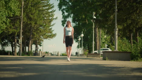 lady walking confidently on interlocked pathway lined with lamp posts, surrounded by tall trees, lush greenery, parked car, and blurred view of person in distance pushing child stroller