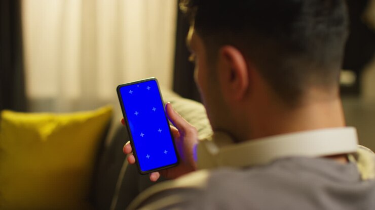 Over The Shoulder Shot Of Man Spending Evening At Home Sitting On Sofa With Wireless Headphones Looking At Blue Screen Mobile Phone