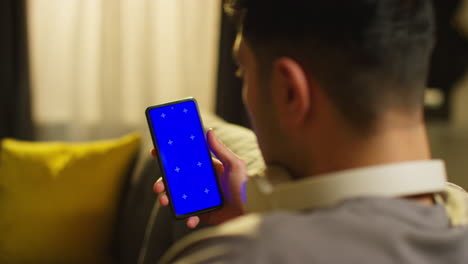 más del hombro del hombre que pasa la noche en casa sentado en el sofá con auriculares inalámbricos mirando la pantalla azul del teléfono móvil