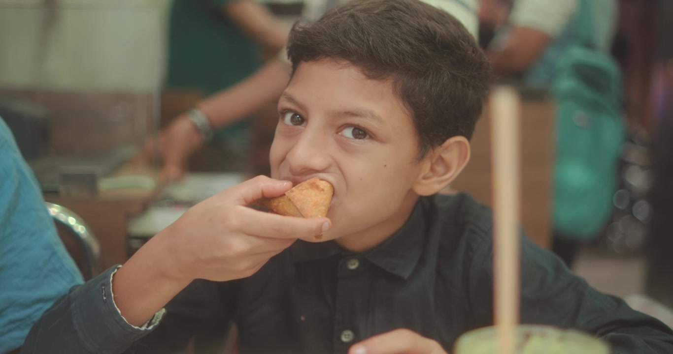 Un giovane ragazzo mangia con gusto una samosa tradizionale, assaporando il delizioso spuntino in un ristorante, celebrando momenti d'infanzia e cucine autentiche.