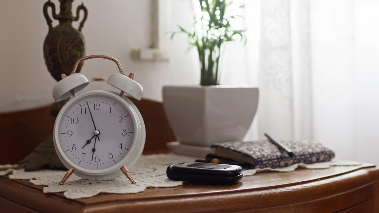 A young woman’s hand reaches to turn off a white analog alarm clock on a bedside table, with soft natural light, a plant, and personal items in the background, close-up shot
