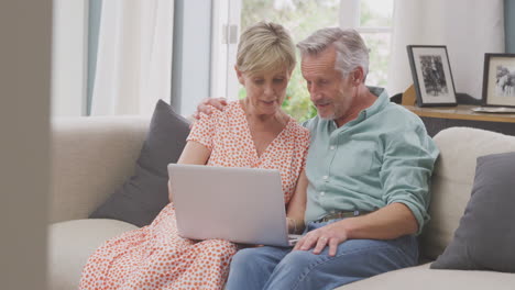 Senior-Retired-Couple-Sitting-On-Sofa-At-Home-Shopping-Or-Booking-Holiday-On-Laptop