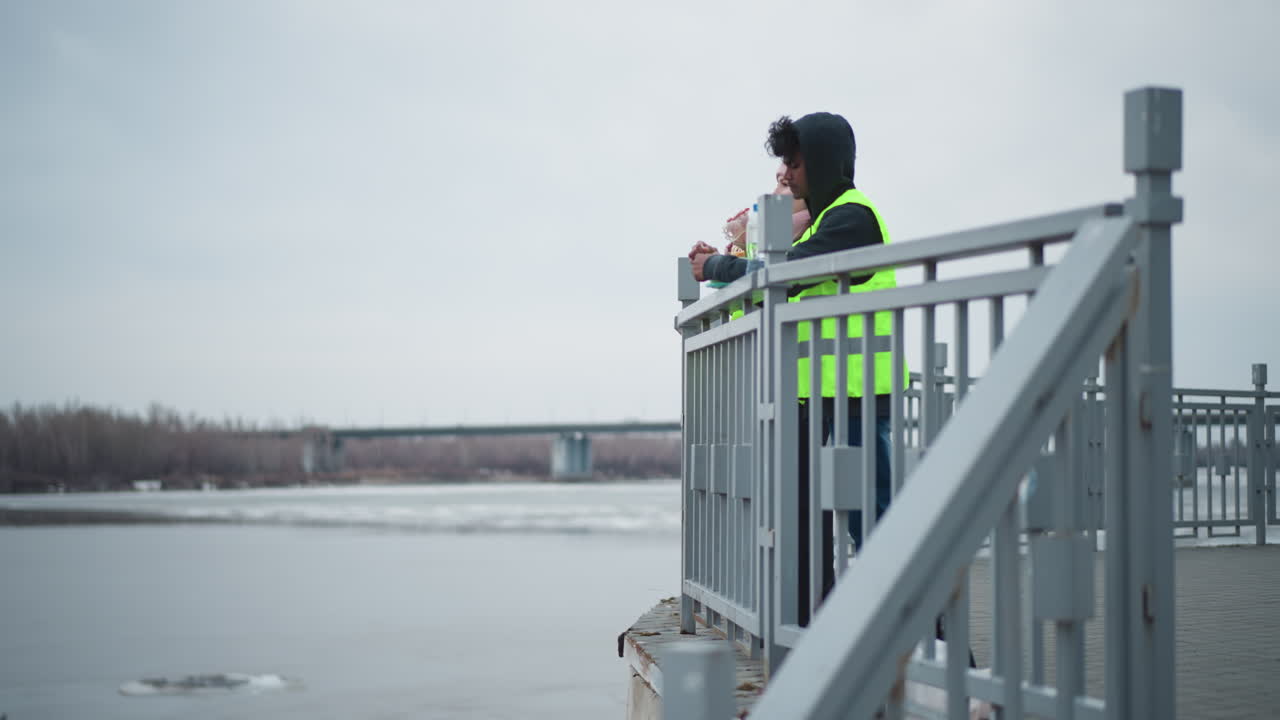 Two people in reflective safety vests stand on riverside promenade near railing eating food in cold weather, with bags on ground, bridge in background, overcast sky, and partially frozen water surface