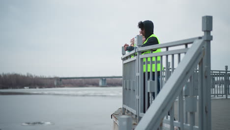 two people in reflective safety vests stand on riverside promenade near railing eating food in cold weather, with bags on ground, bridge in background, overcast sky, and partially frozen water surface
